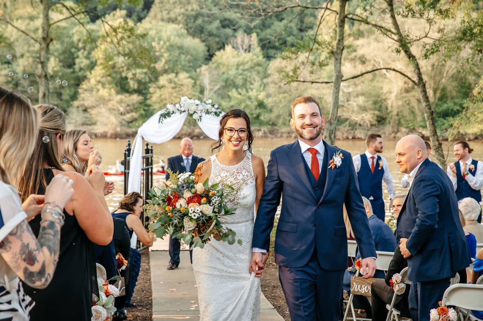 A bride and groom walking back down the aisle past their seated guests after an outdoor ceremony, the bride carrying a bright bouquet.