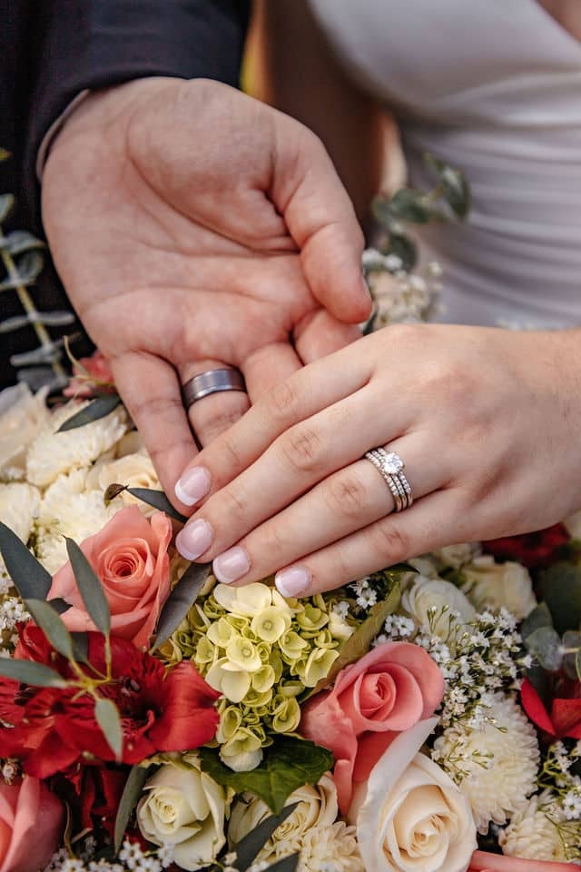 A bride and groom's hands resting on her bouquet, both wedding rings and her engagement ring in close-up.